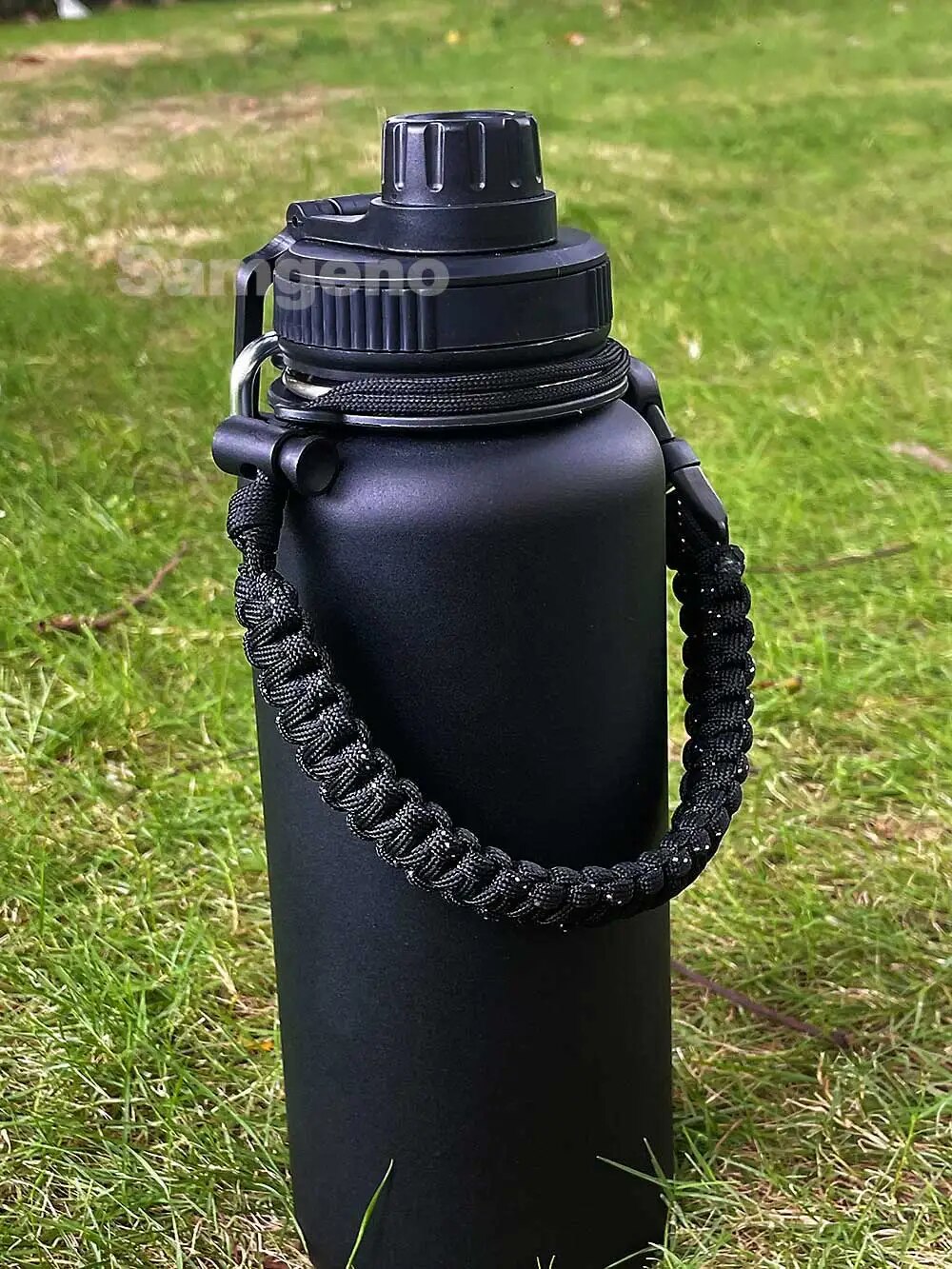 Image for 32 oz or 1000 ML stainless steel water bottle in black color, with double wall vacuum insulation. The bottle has dual lid with built-in carrying handle. The image shows the bottle is lying on the grass and has a black carry strap attached to bottle's neck below the lid.