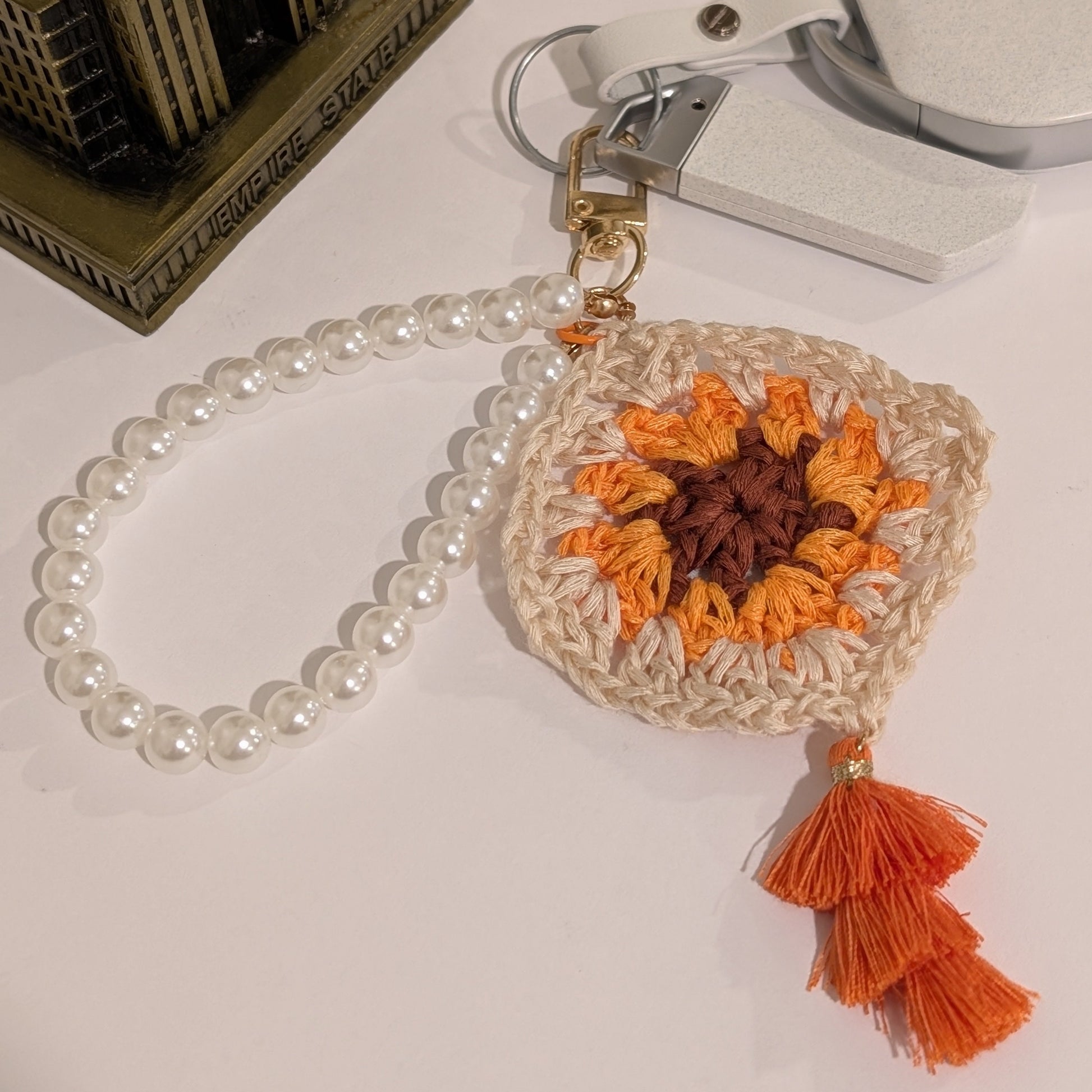 Beige crocheted keychain with orange and brown flower design, and orange tassel on a white surface