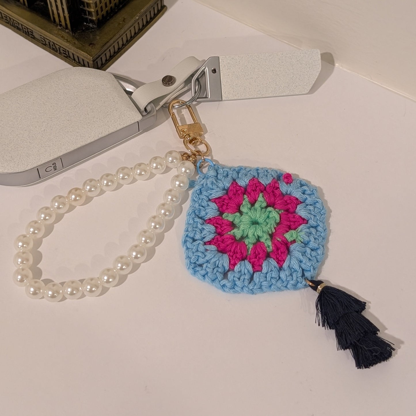 Blue Crocheted keychain with colorful design, white pearl bracelet and black tassel on a white surface.