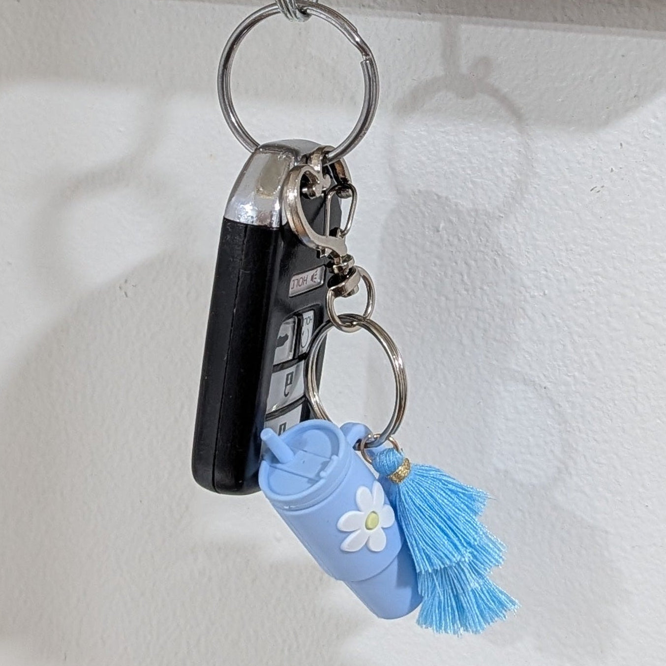 Blue little silicone tumbler key chain with crochet flower hanging on wooden key holder with a glass jar and decorative items on a white wall.