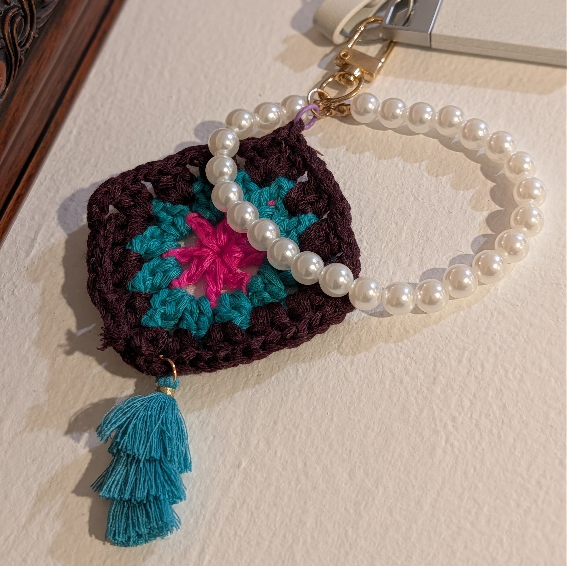 Dark brown crocheted keychain with colorful flower design, teal tassel and pearl chain on a beige surface