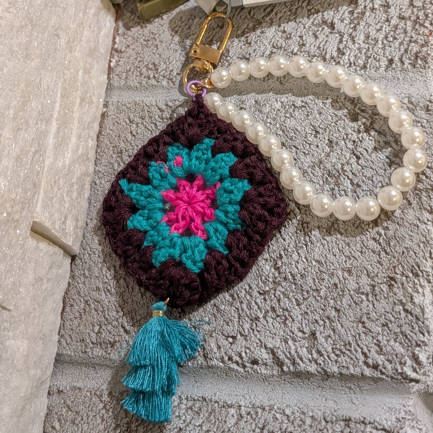 Dark brown crocheted keychain with colorful flower design, teal tassel and pearl chain on a gray stone surface