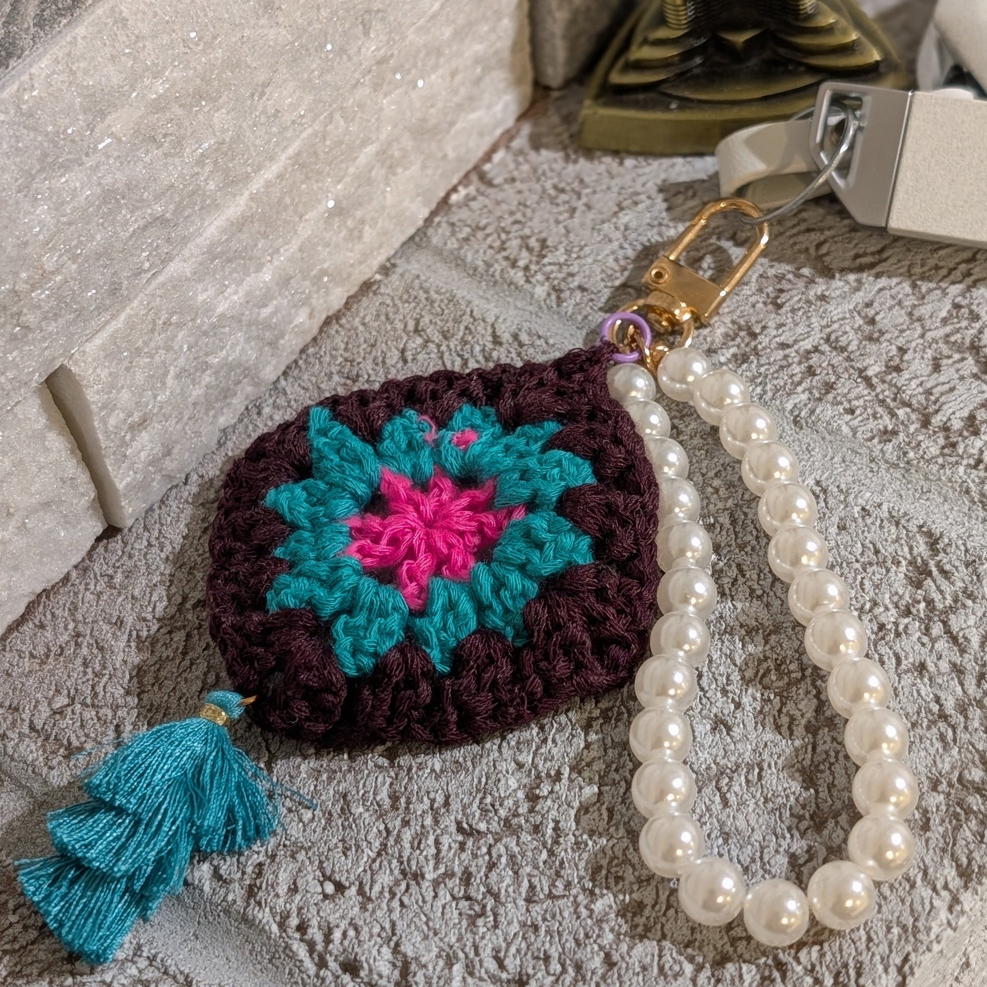 Dark brown crocheted keychain with colorful flower design, teal tassel and pearl chain on a stone surface