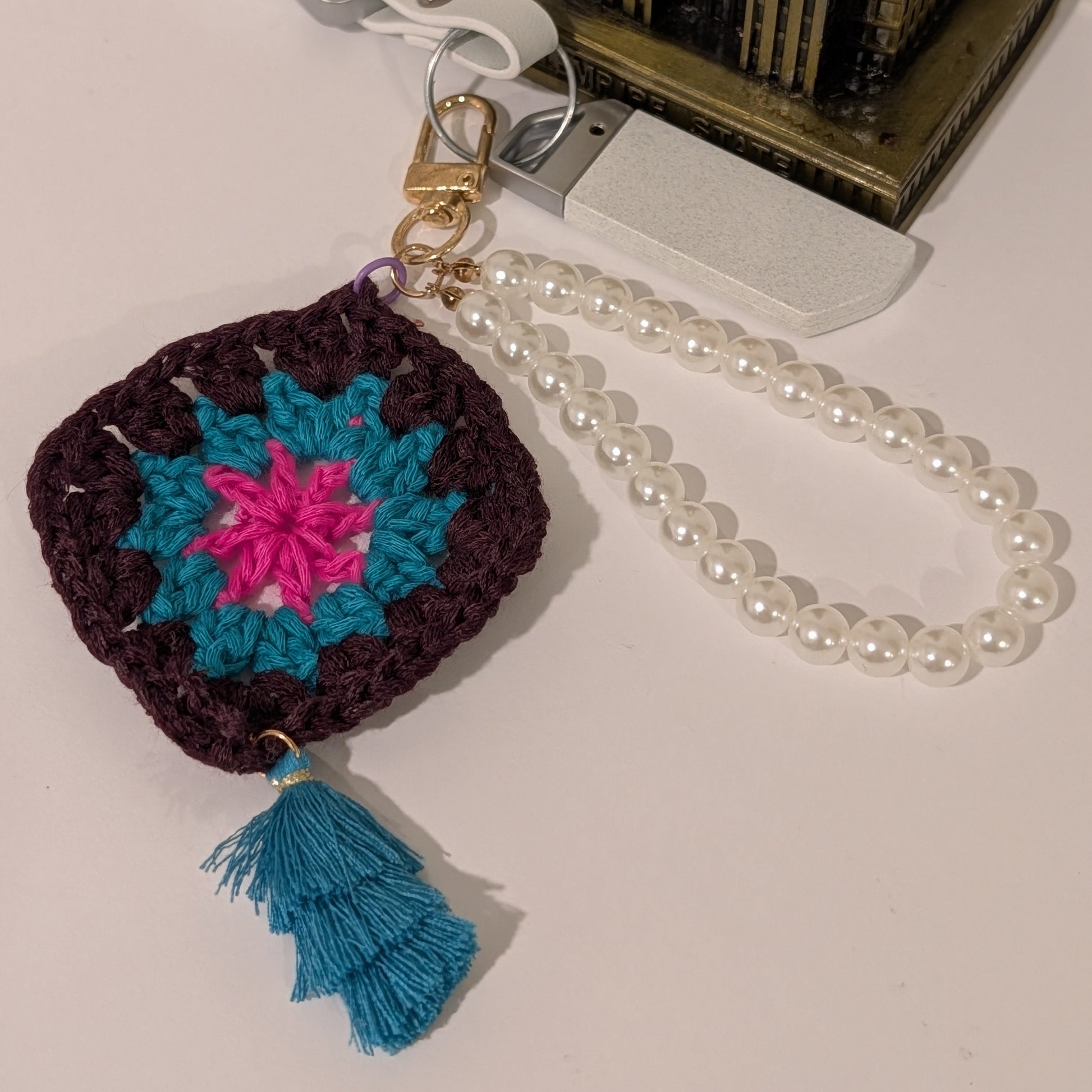 Dark brown crocheted keychain with colorful flower design, teal tassel and pearl chain on a white surface