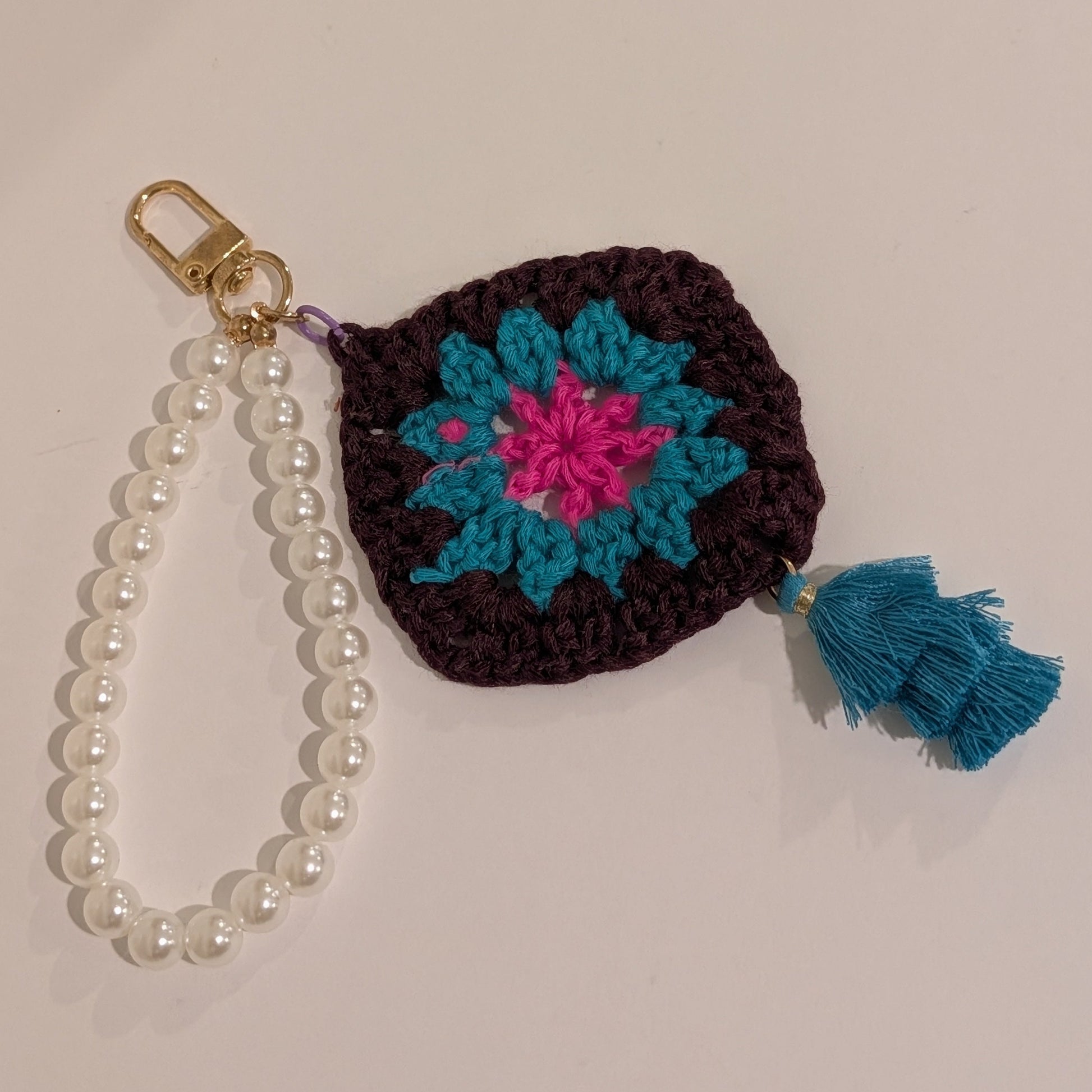 Dark brown crocheted keychain with floral design, pearl chain, and teal tassel on a light background