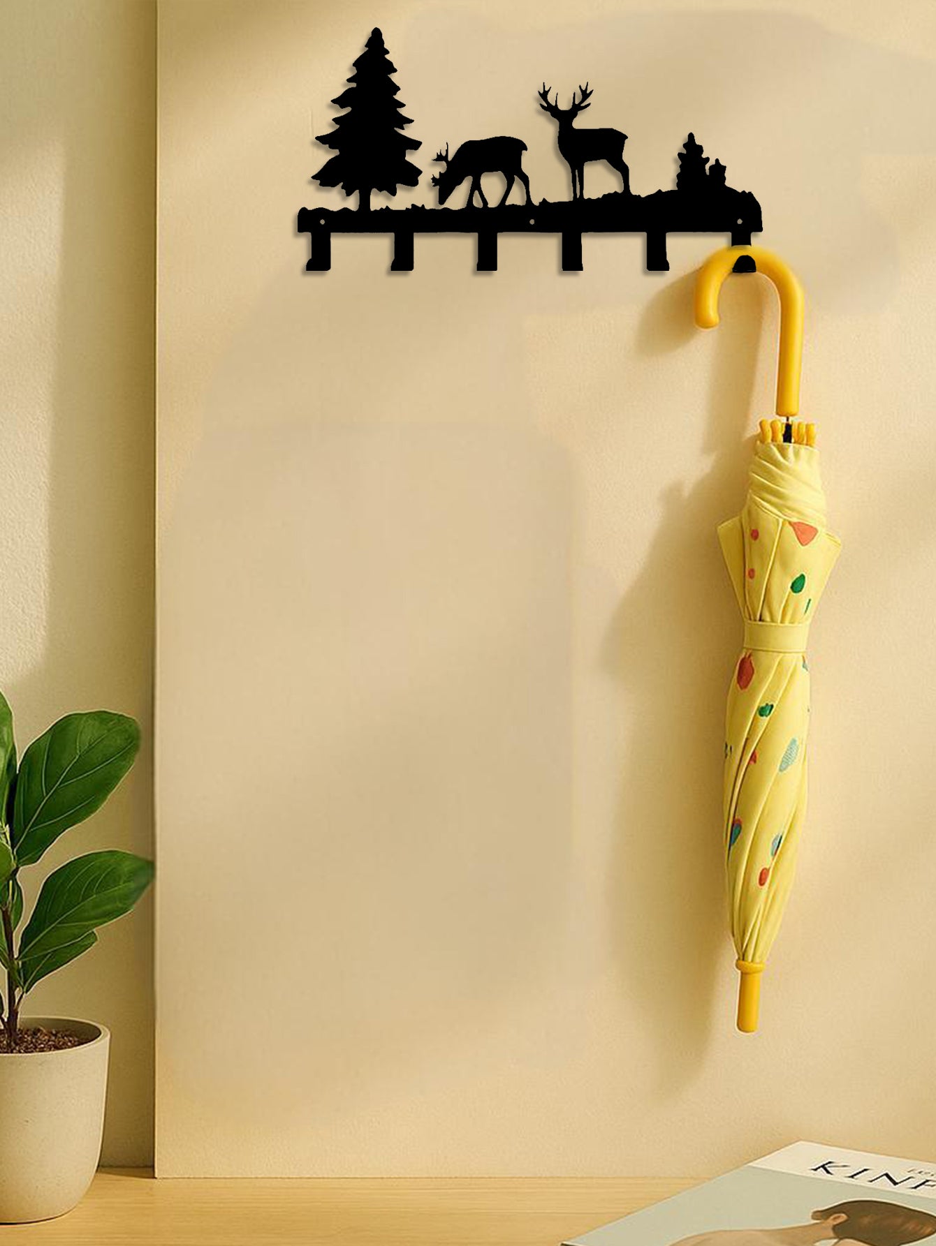 Decorative 6-hook wall mounted key and coat rack with elk and tree designs holding a yellow umbrella against a beige wall.