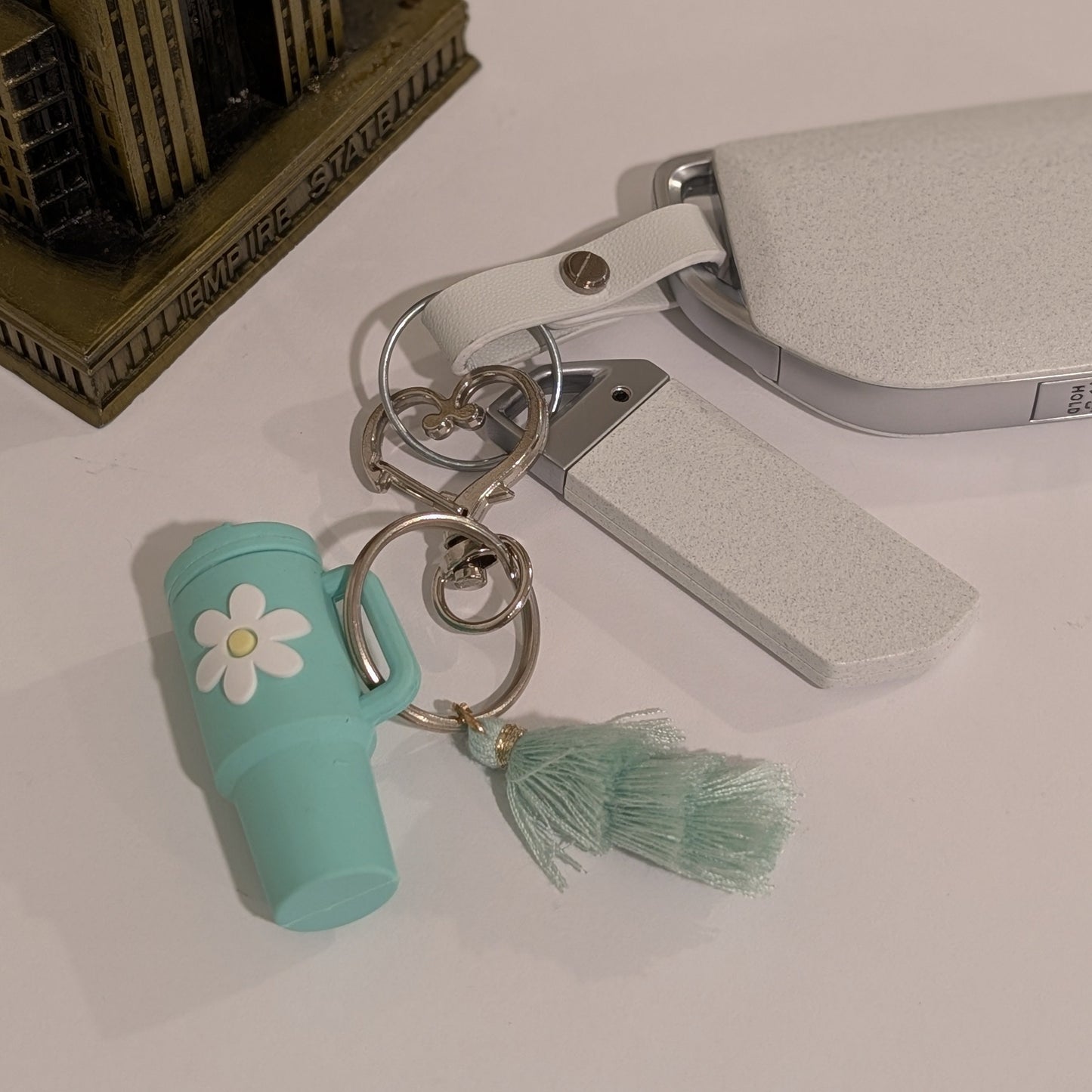 Green tumbler cup-shaped keychain with a flower and tassel attached to a car key on a light white background