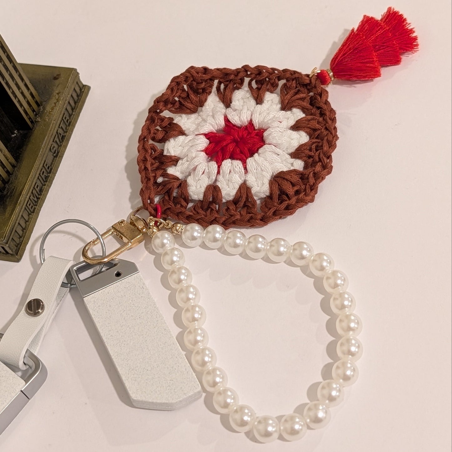 Maroon crocheted square keychain with red center and white border, attached to a pearl bracelet on a light surface.