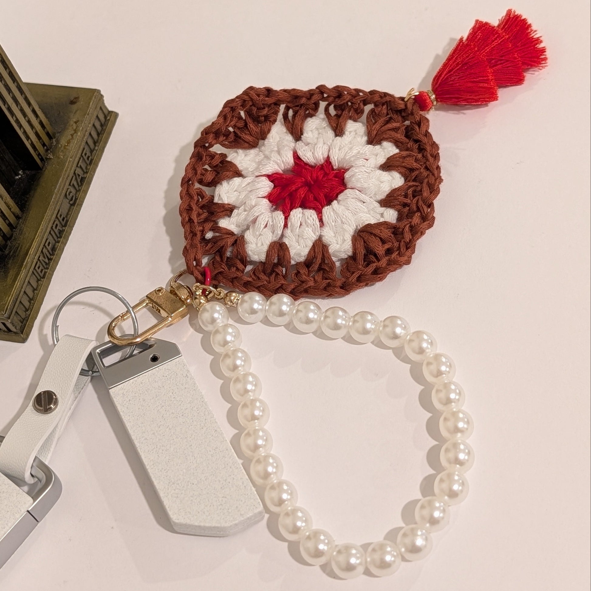 Maroon crocheted square keychain with red center and white border, attached to a pearl bracelet on a light surface.