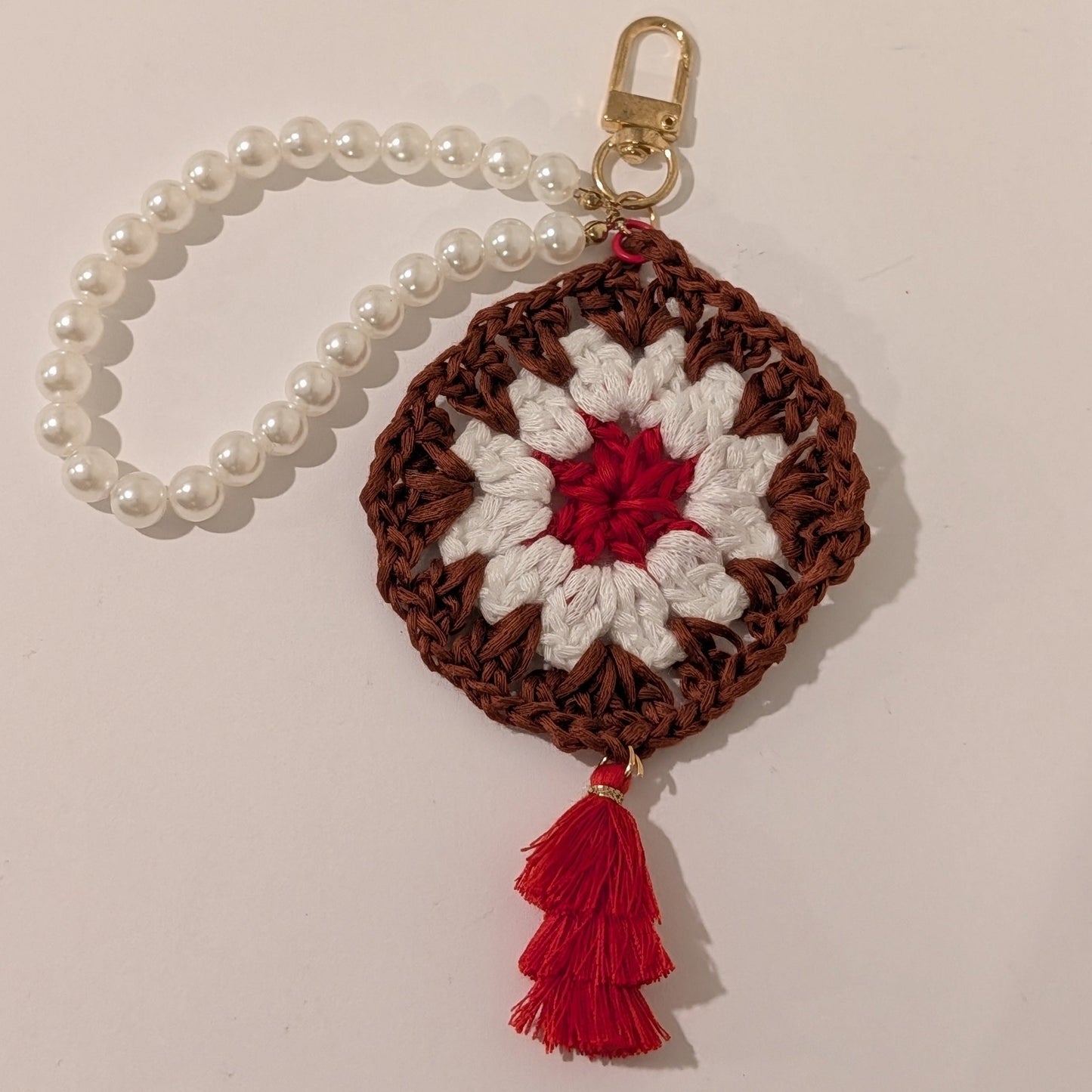Maroon crocheted square keychain with red center and white border, attached to a pearl bracelet on a white surface.
