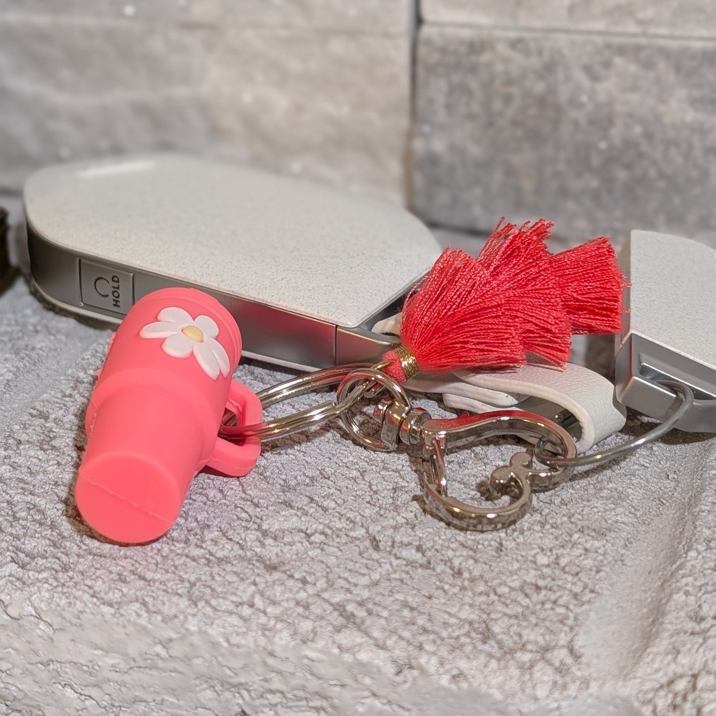 Pink silicone tumbler keychain with a flower charm, heart shaped buckle and red tassel attached to a car key on a gray background