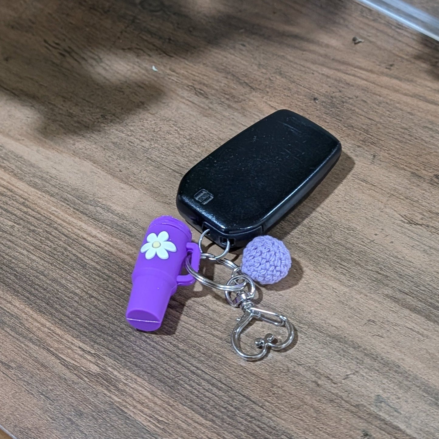 Purple little silicone tumbler keychain with daisy flower design and crochet ball on a wooden surface with plants in the background