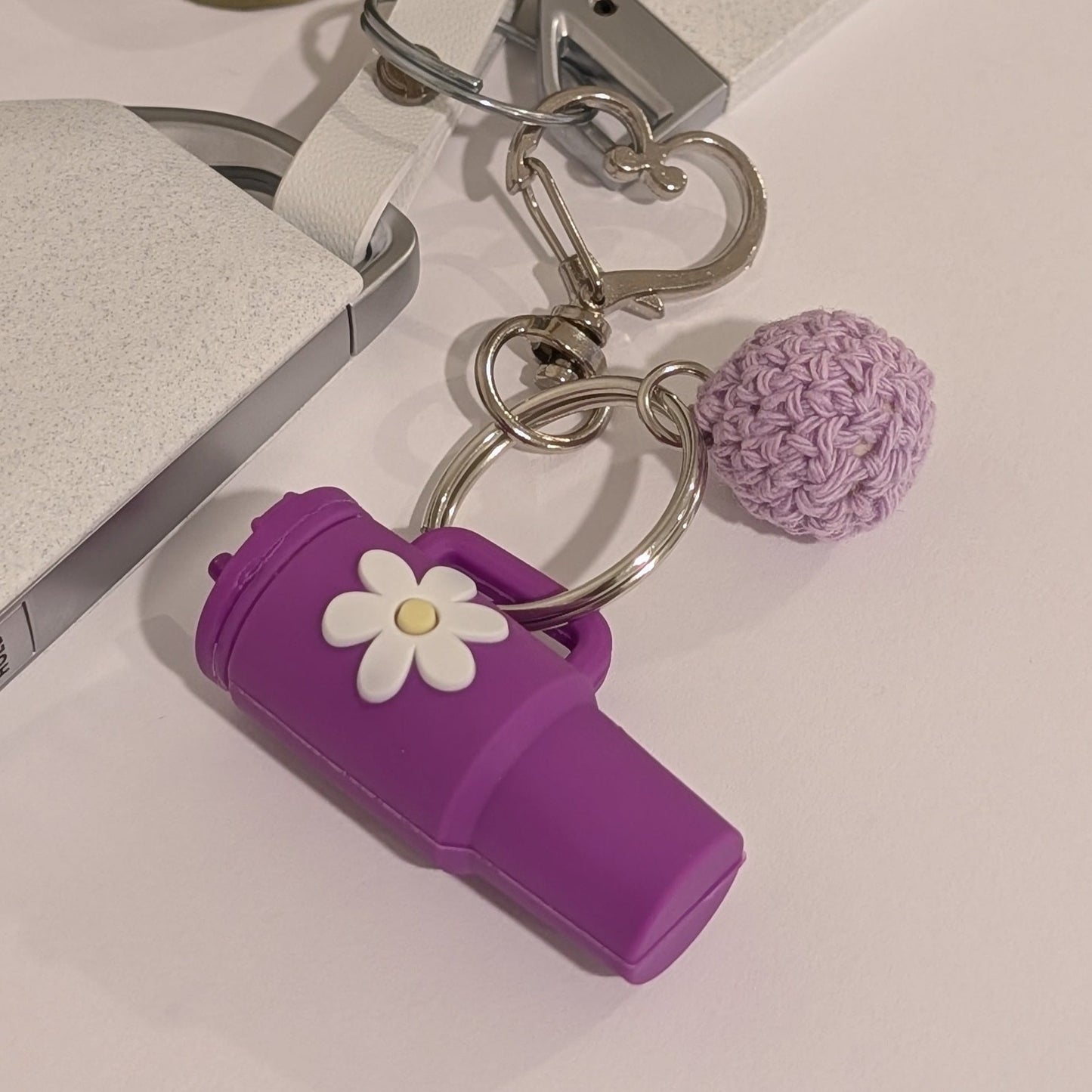 Purple silicone Keychain with black tumbler, flower charm, and pom-pom on a light surface.