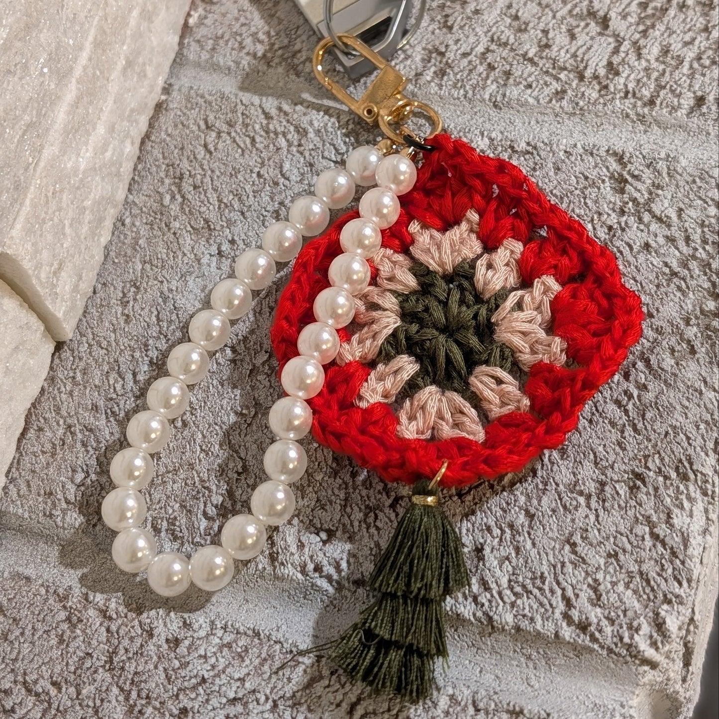 Red crocheted keychain with moss color tassel and white pearl chain bracelet on stone surface