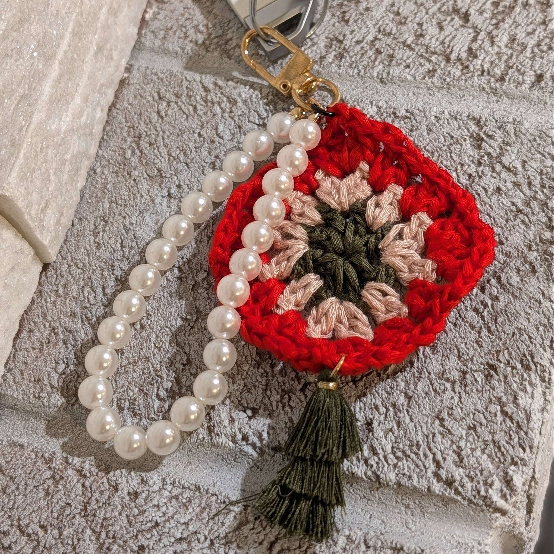 Red crocheted keychain with moss color tassel and white pearl chain bracelet on stone surface