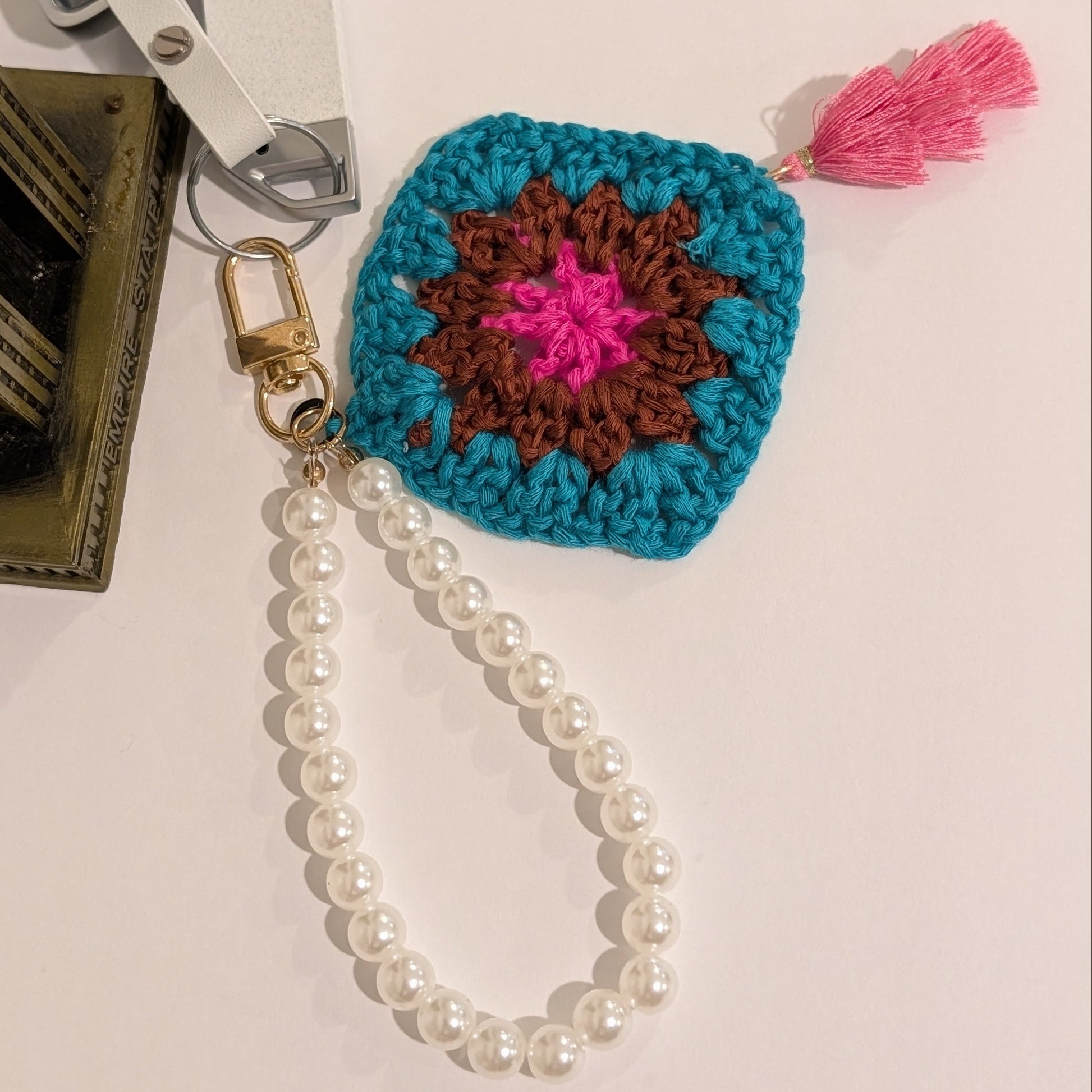 Teal crocheted keychain with colorful design and pearl chain bracelet on a neutral background