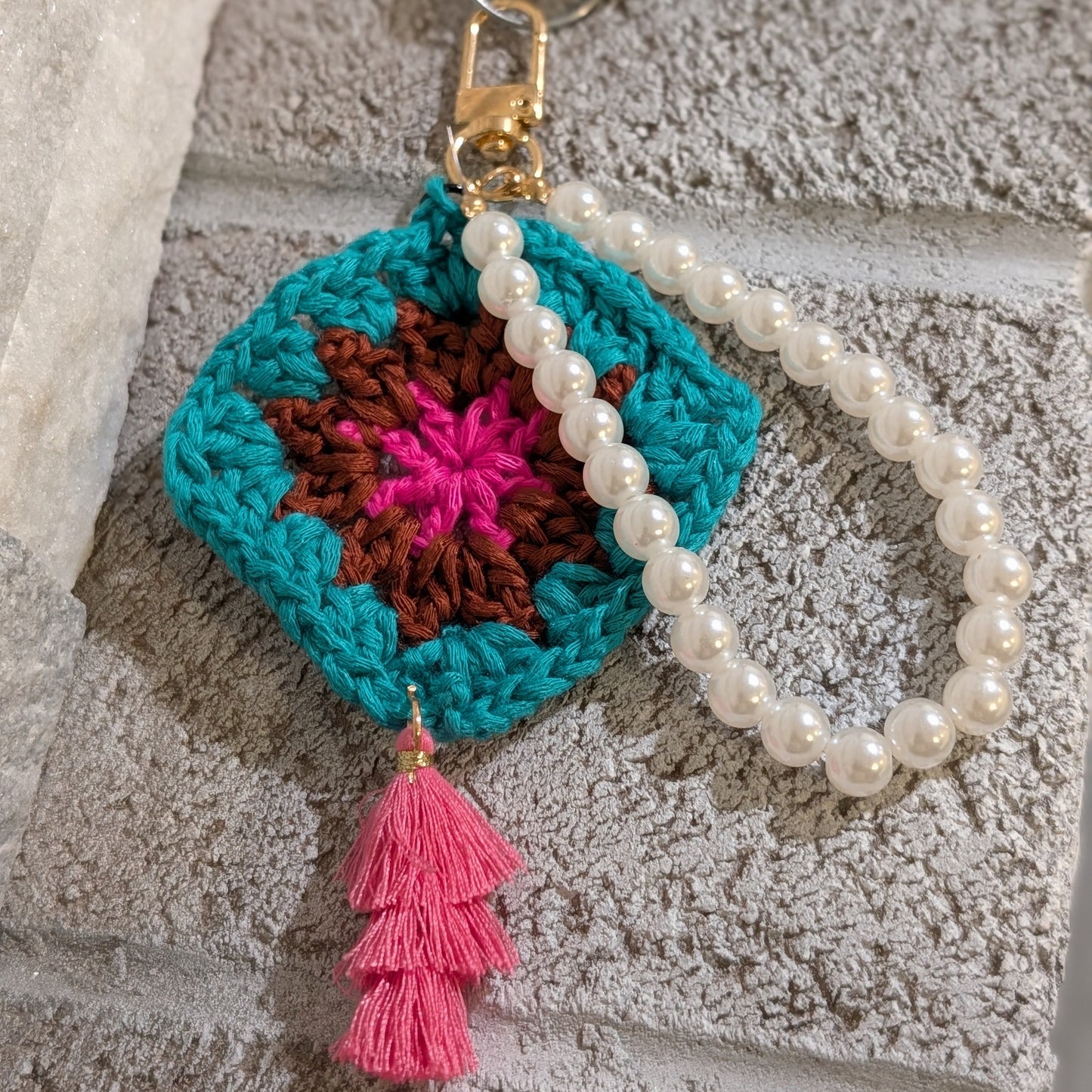 Teal crocheted keychain with colorful design, pink tassel and white beaded bracelet on a textured surface