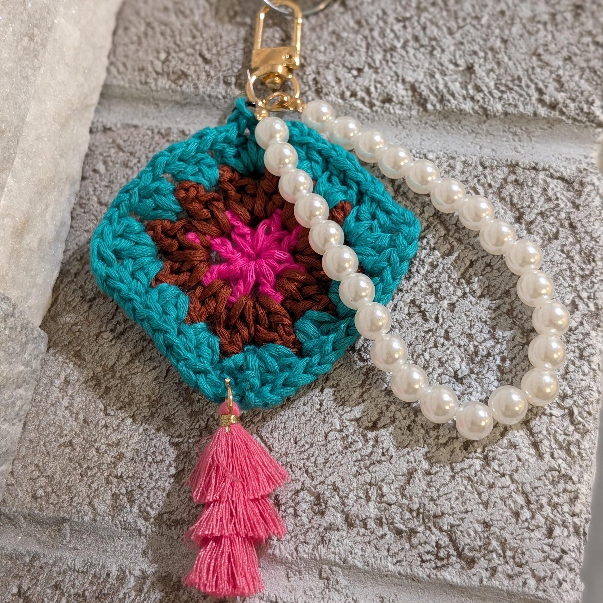 Teal crocheted keychain with colorful design, pink tassel and white beaded bracelet on a textured surface