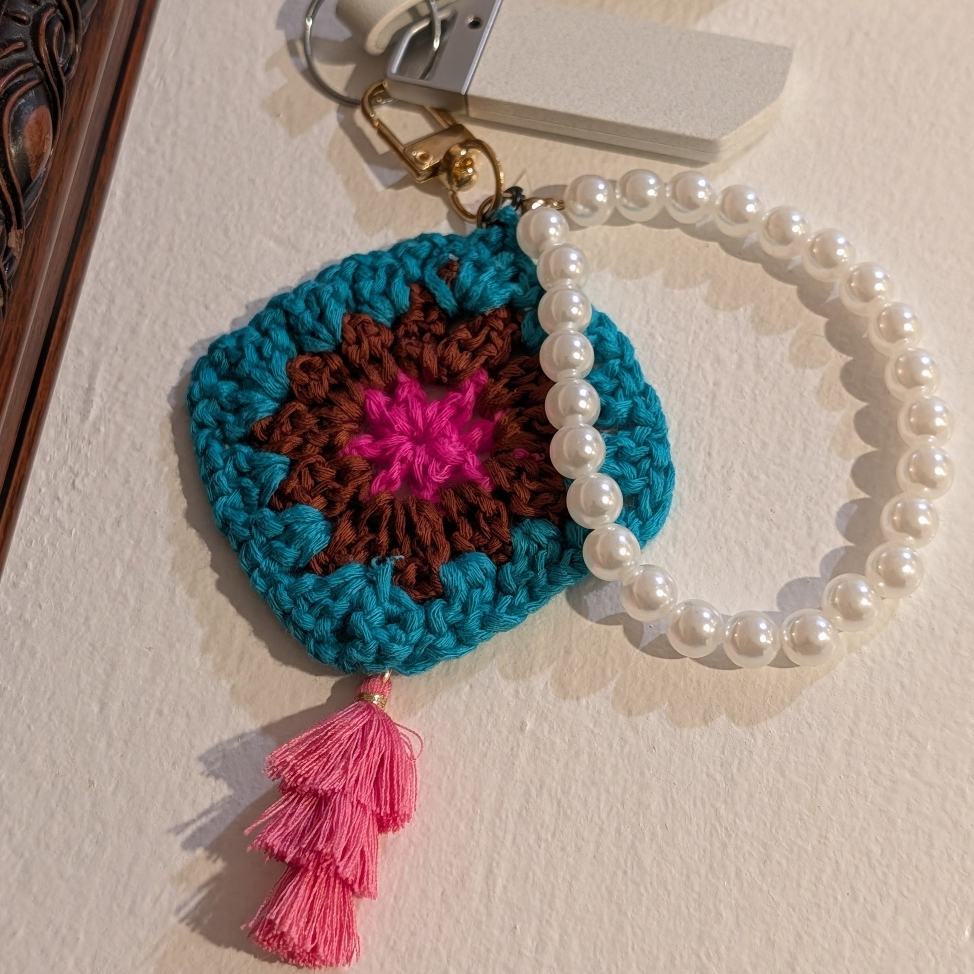 Teal crocheted keychain with pink tassel and pearl bracelet on a beige surface