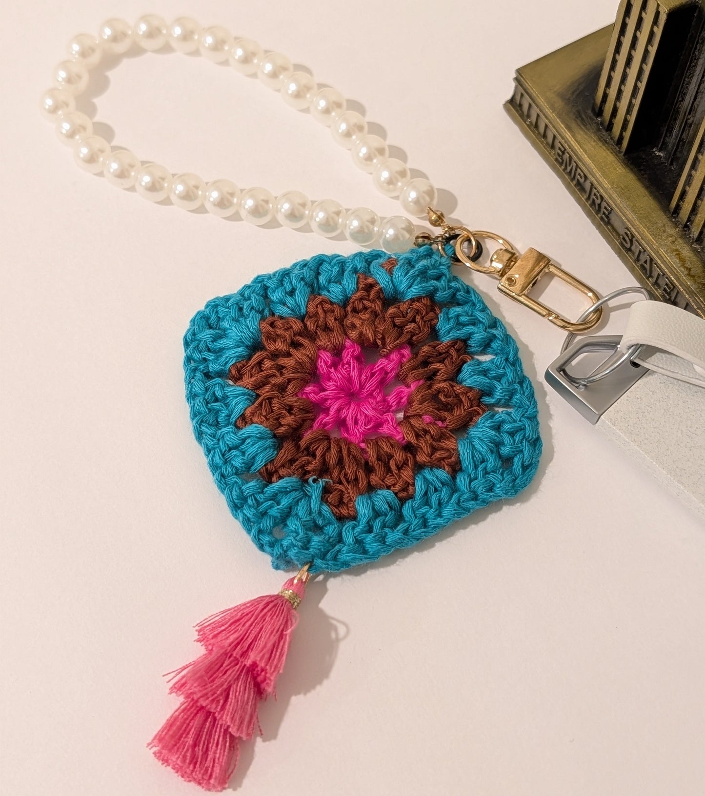 Teal crocheted square keychain with pink tassel and pearl bracelet on a light background