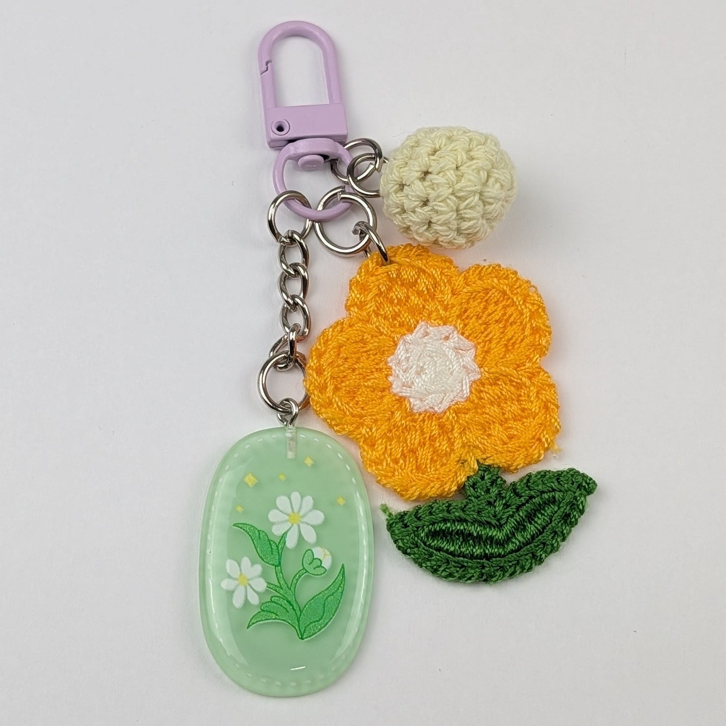 Yellow crocheted flower keychain with a beige crochet ball, green leaf, and a green resin tag with a floral design on a light gray background.