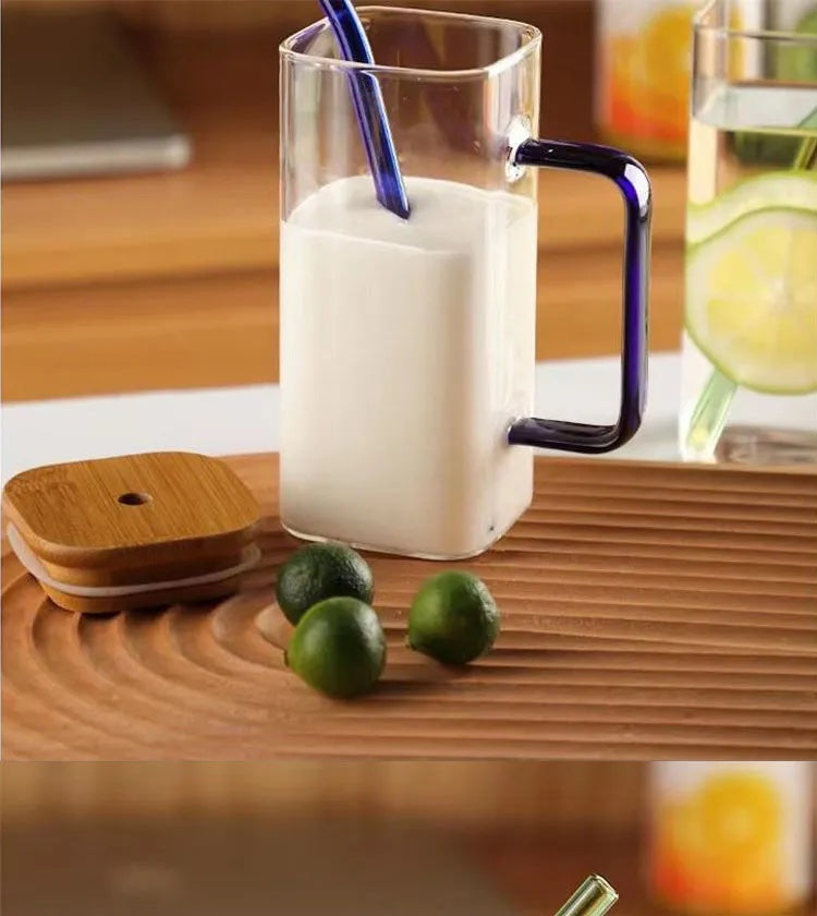 Image with a square glass mug with blue handle and straw. The wooden lid is placed on the side. The mug is placed on a wooden surface with three whole green lemons placed next to it. The mug has milk in it.