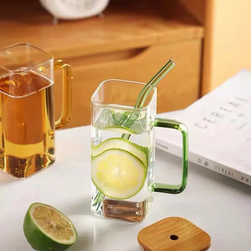 Image with two square glass mugs placed on a white flat surface with a wooden lid and a lemon cut placed on the side. The mug on the right side has green handle and straw without lid on it. The other mug has transparent handle without straw.  A partially visible book is placed next to both mugs