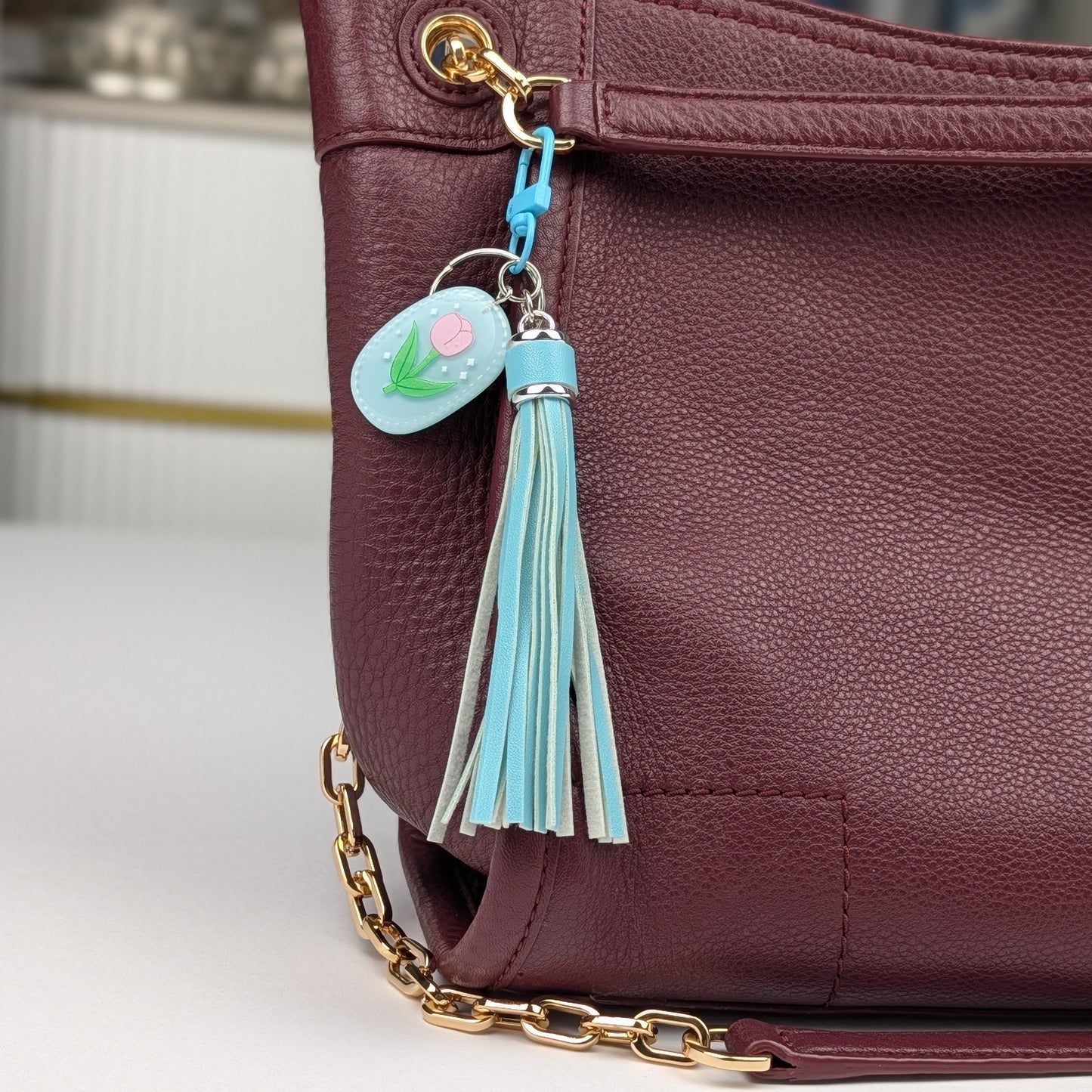 Blue tassel keychain and tulip charm tag attached to a Maroon handbag on a light gray background
