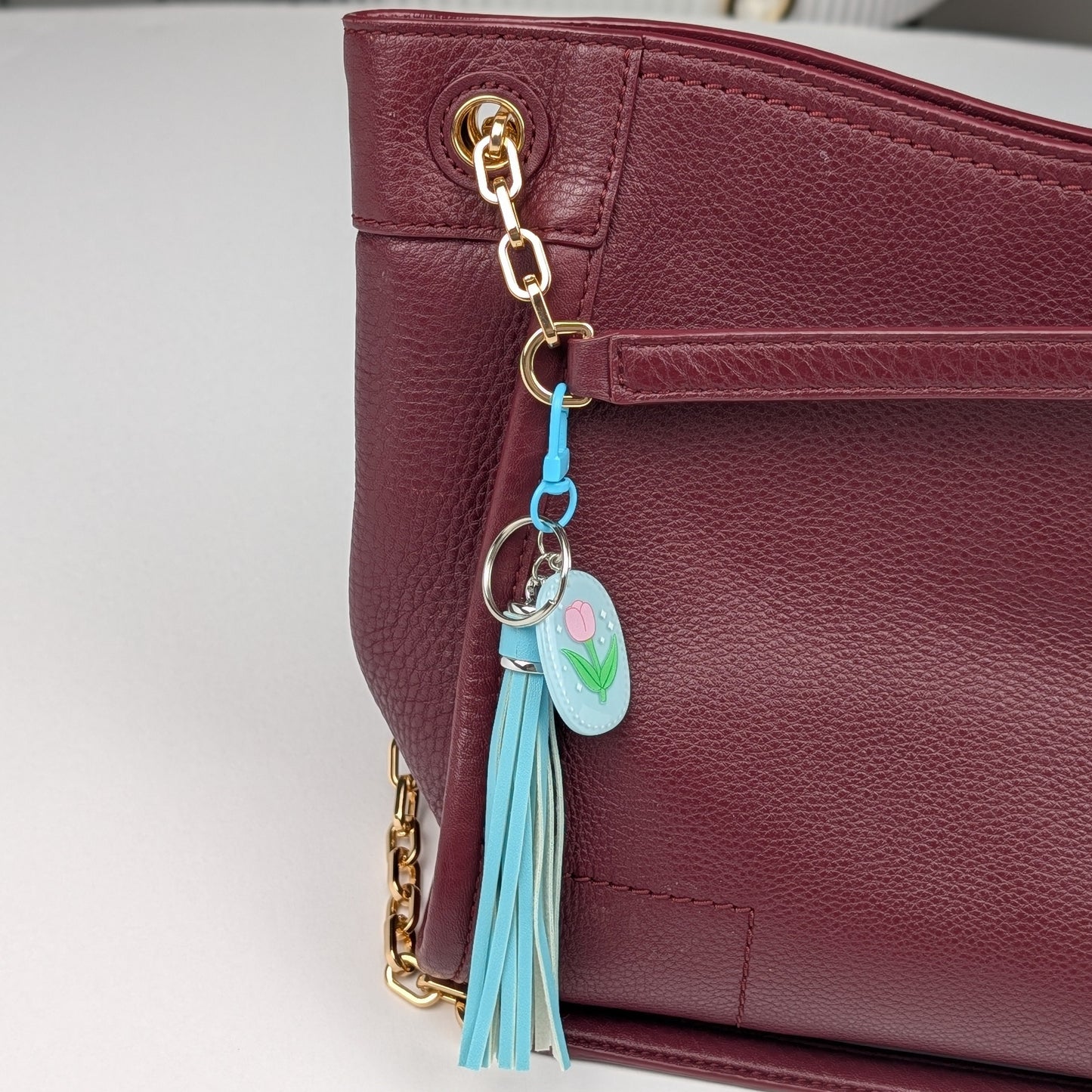 Blue tassel keychain attached to a Maroon handbag on a light gray background
