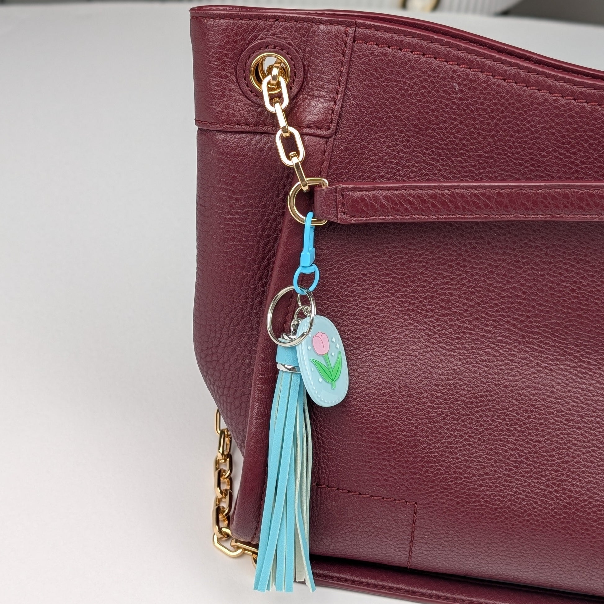 Blue tassel keychain attached to a Maroon handbag on a light gray background
