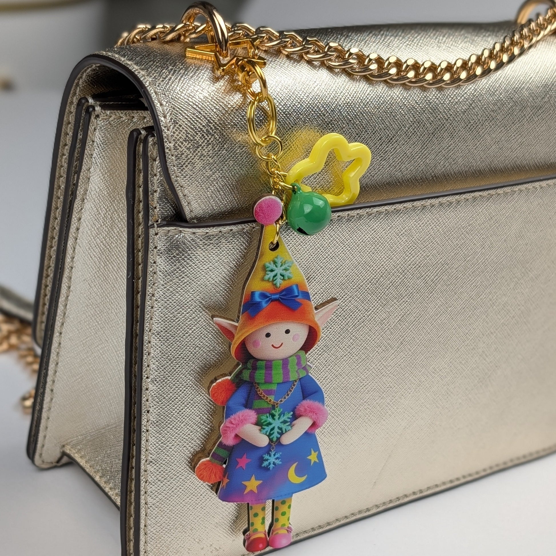 Colorful holiday themed fairy doll keychain with golden star, orange flower, and dark green bell charm attached to a golden women handbag on a white background