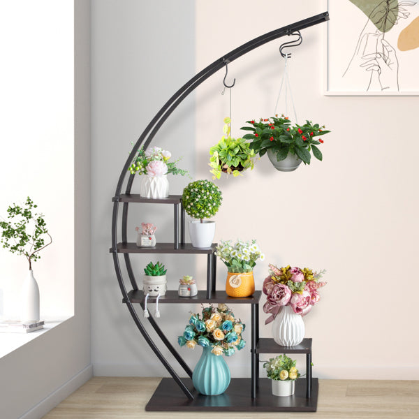 Curved black metal 5 layer plant stand with various potted plants and flowers against a light wall.