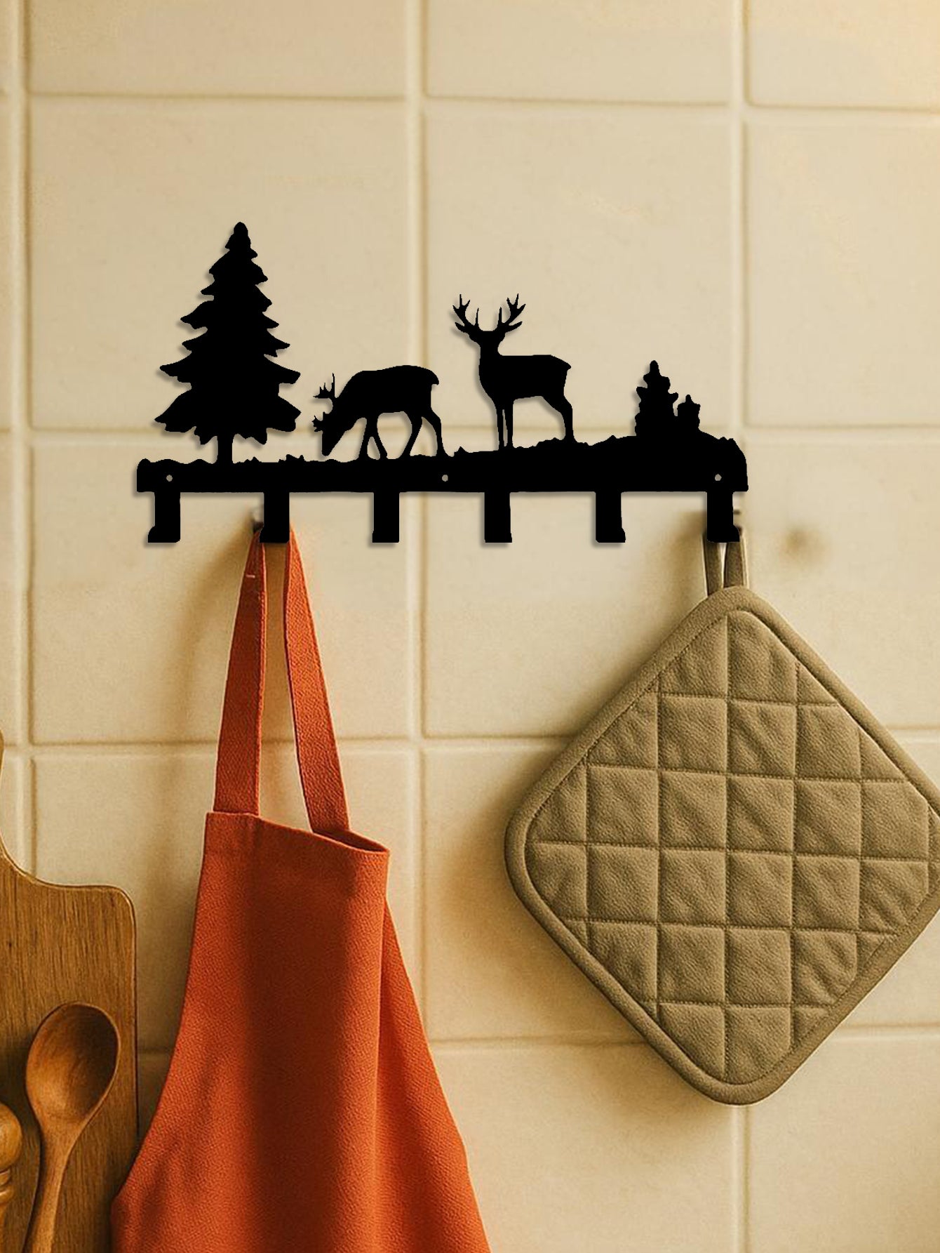 Decorative 6-hook wall mounted key and coat rack with deer and tree design on a tiled wall, with an apron and pot holder hanging from it.