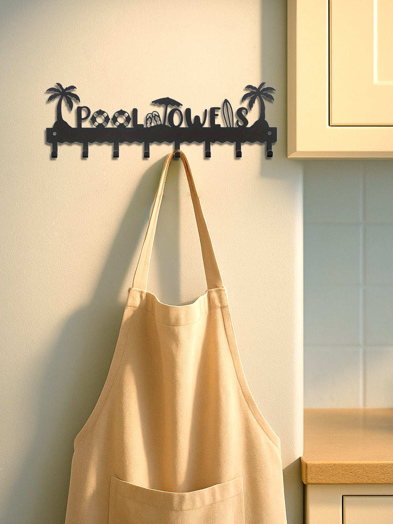 Decorative wall hook with 'Pool Towels' text hanging an apron on a kitchen wall.