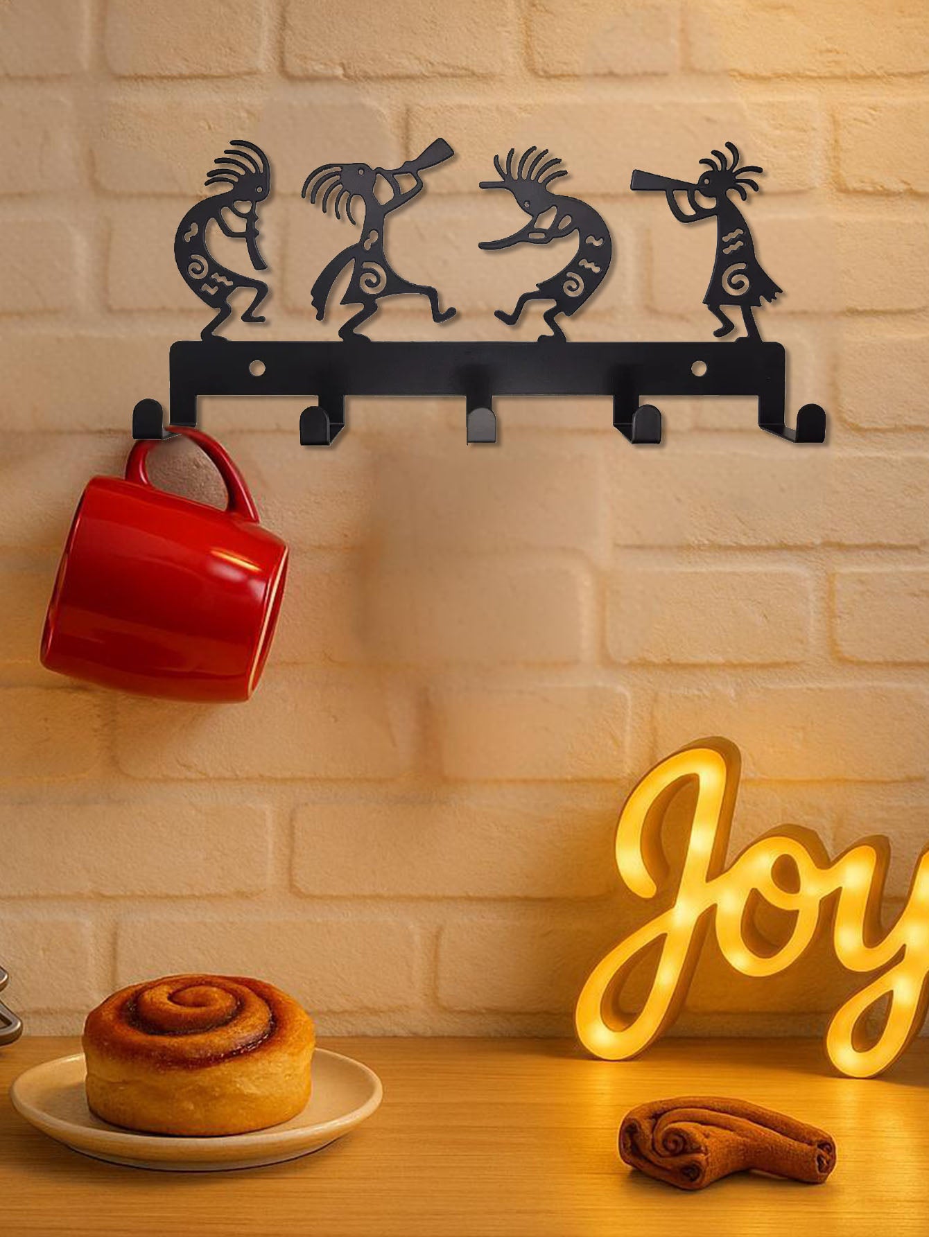 Decorative wall hook with four hooks, a red mug, cinnamon roll, and 'Joy' sign on a wooden surface.