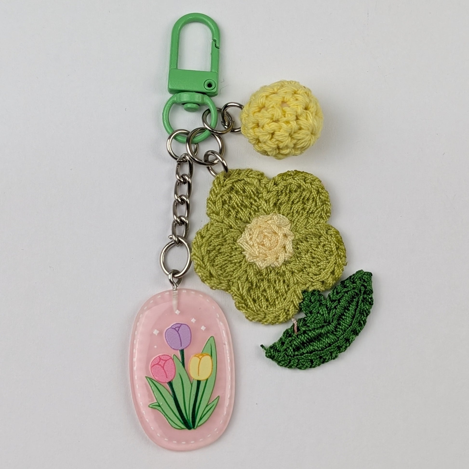 Floral keychain with a light green crochet flower with green leaf, light yellow crochet ball and pink resin floral tag on a light gray background