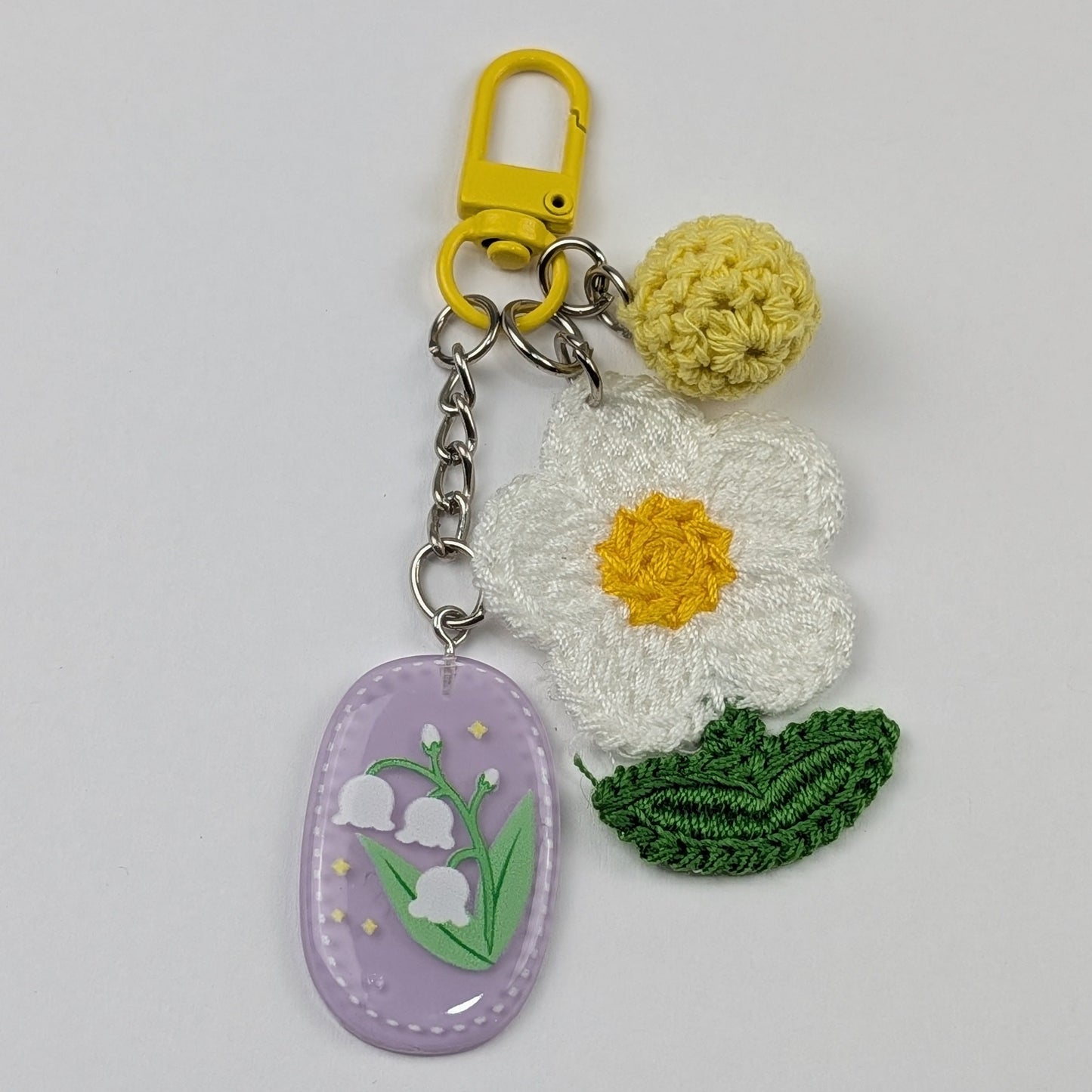 Floral keychain with a white crochet flower with green leaf, light yellow crochet ball and purple resin floral tag on a light gray background