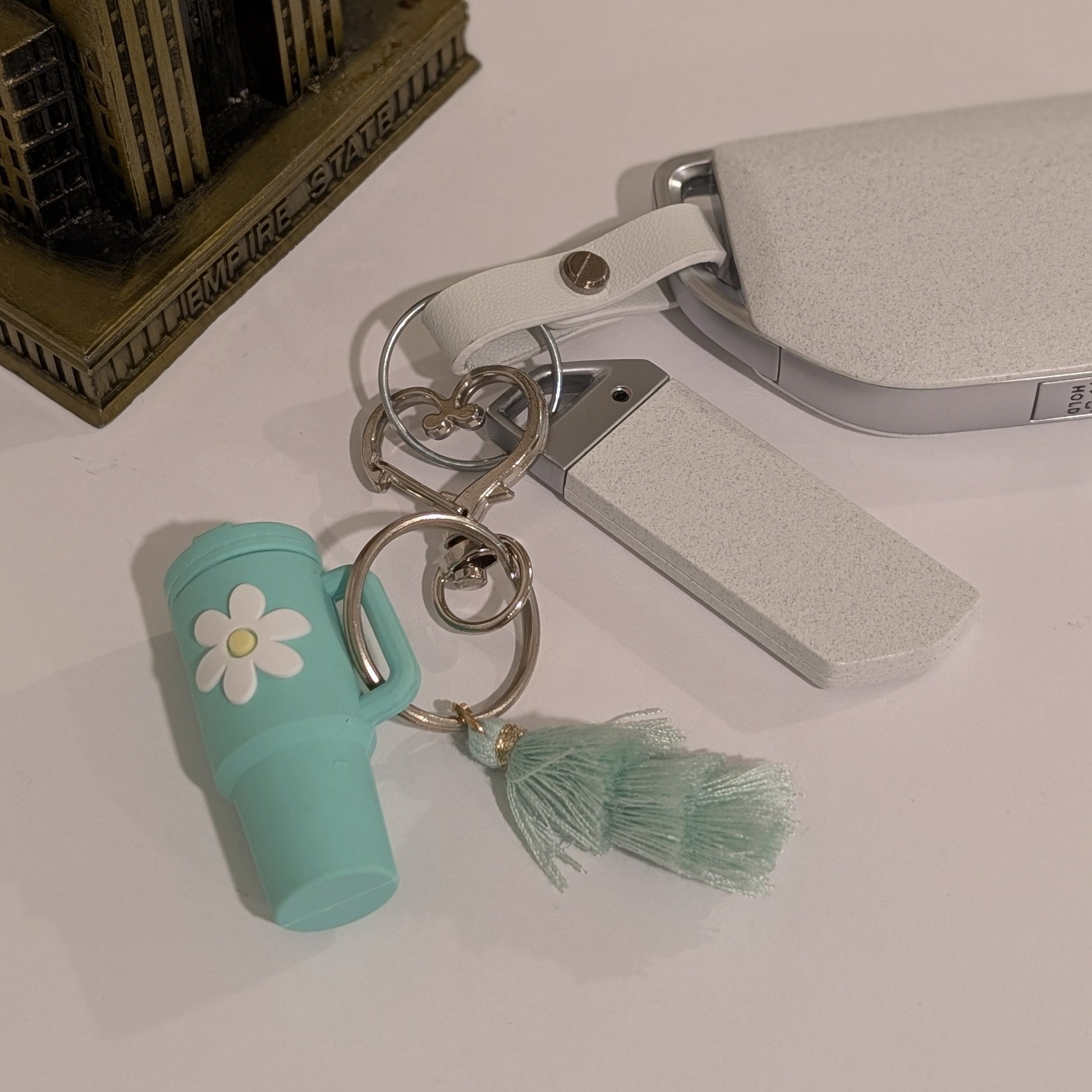 Green tumbler cup-shaped keychain with a flower and tassel attached to a car key on a light white background