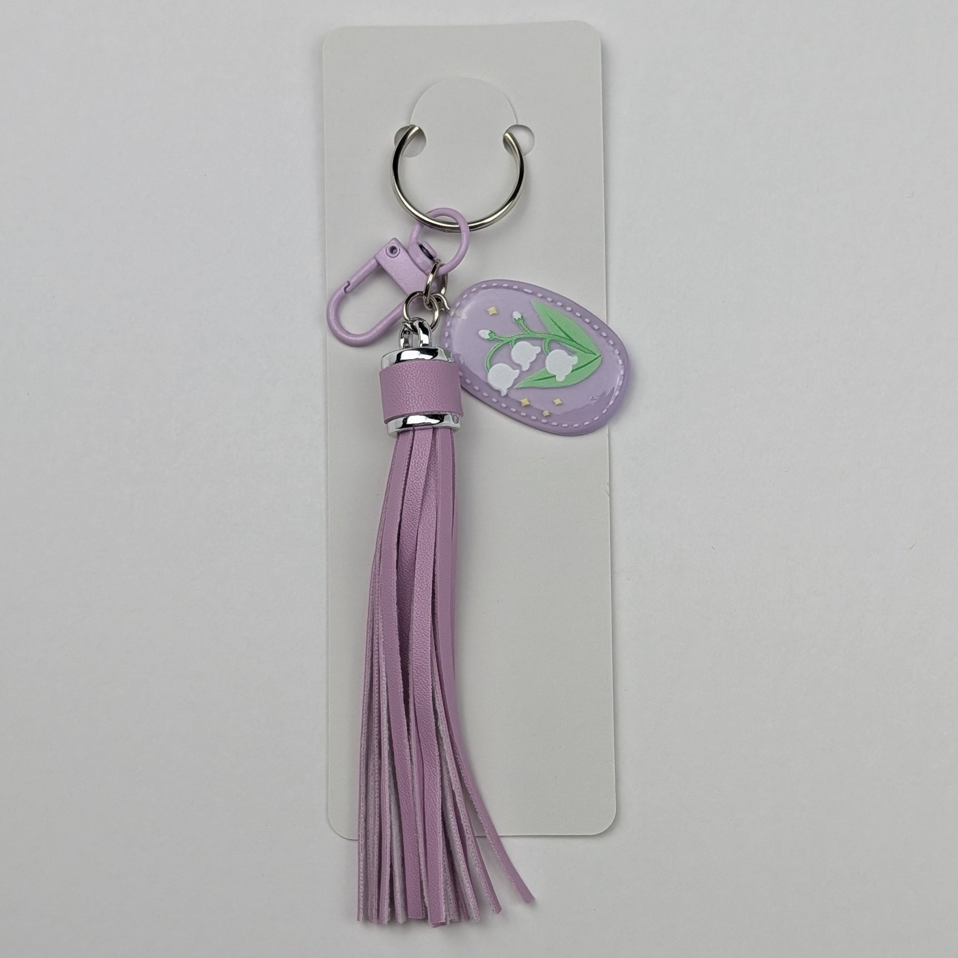 Purple faux leather tassel keychain with a floral tag attached to a packaging card on a clear background