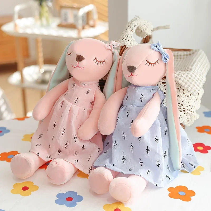 Two bunny plush toys, one pink and one blue, sitting on a floral-patterned surface.