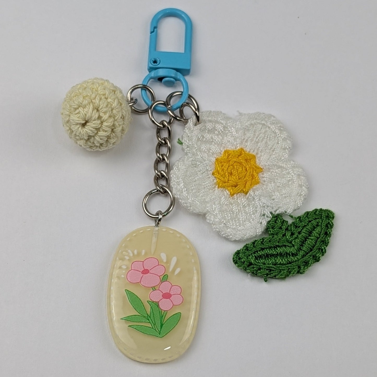White crocheted flower keychain with a light yellow crochet ball, green leaf, and a beige resin tag with a floral design on a light gray background.