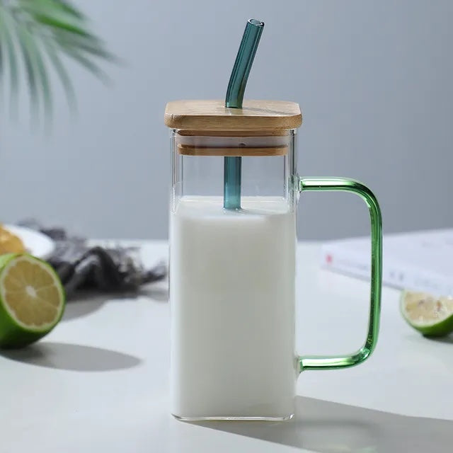 Image with a square glass mug with green handle and straw. It has a wooden lid placed on top with straw inserted through it. The mug is placed on a flat white surface with a sliced green lemon on the side. It has milk in it.