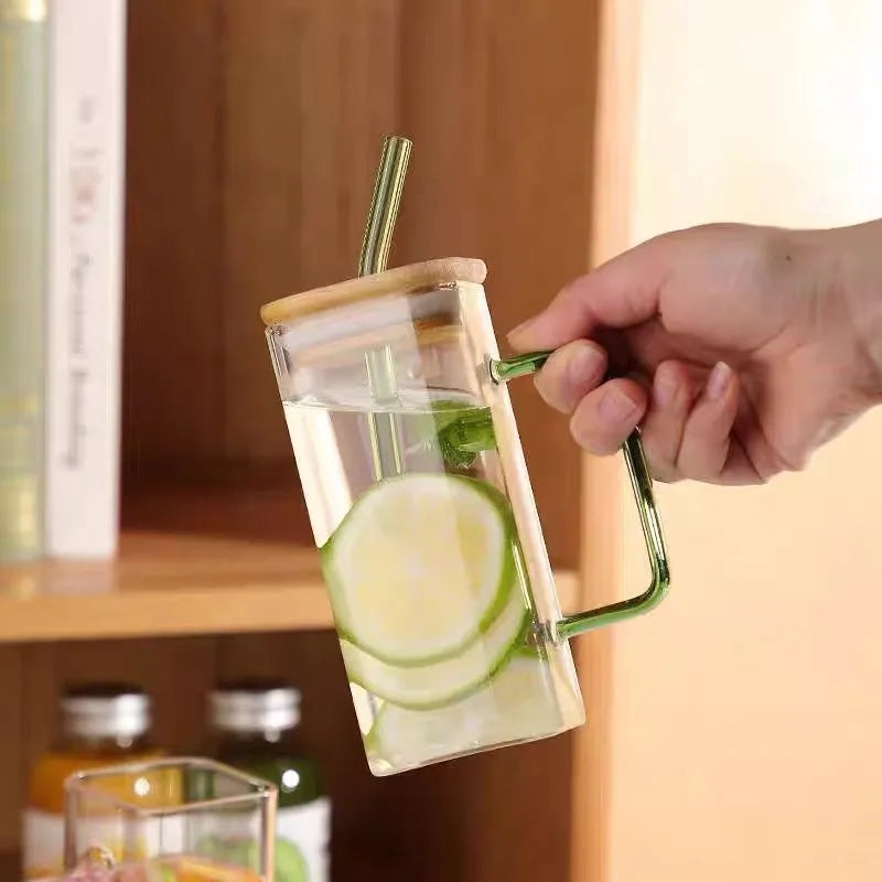 Image with a square glass mug held in a person's hand. It has water and lemon slices inside. The handle and straw color is green. The straw is inserted through the wooden lid.