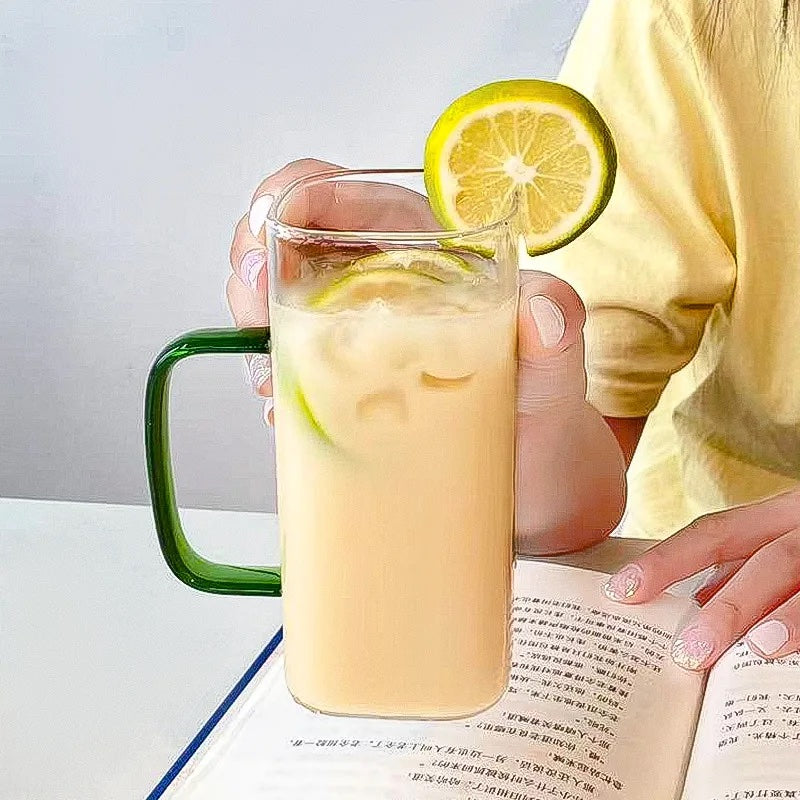 Image with a square glass mug with green handle without lid and straw. It has white drink and with lemon slices inside. A lemon slice is cut-placed on the edge of the mug at the top. The mug is shown held in a person's hand hovering over a book.