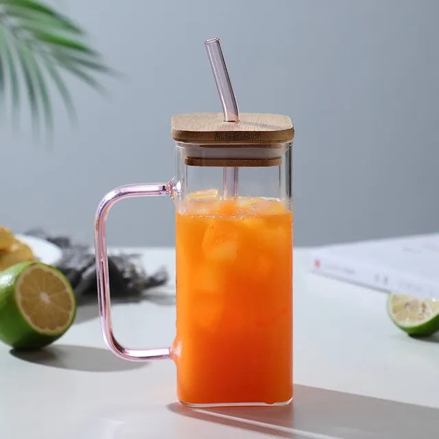 Image with a square glass mug with pink handle and straw. It has a wooden lid placed on top with straw inserted through it. The mug is placed on a flat white surface with a sliced green lemon on the side. It has orange colored drink in it.