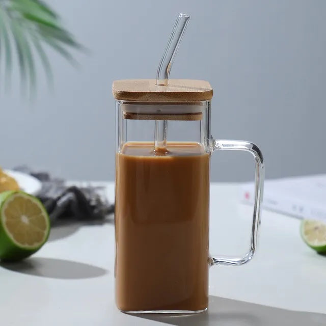 Image with a square glass mug with transparent handle and straw. It has a wooden lid placed on top with straw inserted through it. The mug is placed on a flat white surface with a sliced green lemon on the side. It has coffee in it.
