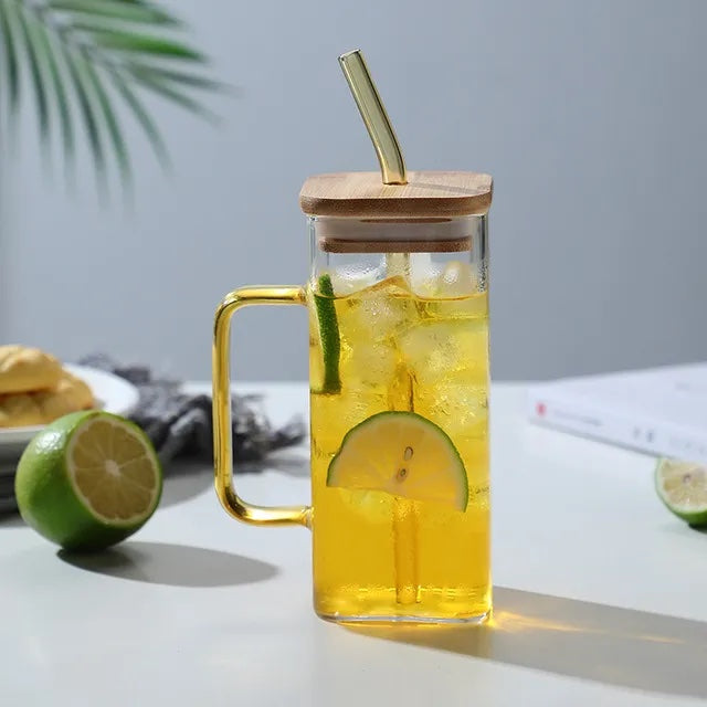 Image with a square glass mug with yellow handle and straw. It has a wooden lid placed on top with straw inserted through it. The mug is placed on a flat white surface with a sliced green lemon on the side. It has yellow colored drink in it with lemon slides inside.