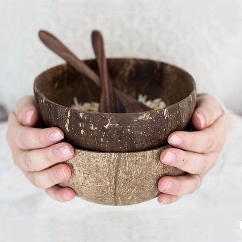 Image for person holding two coconut bowls one on top of the other having two wooden spoons in it. Only person's hands and bowls are visible.
