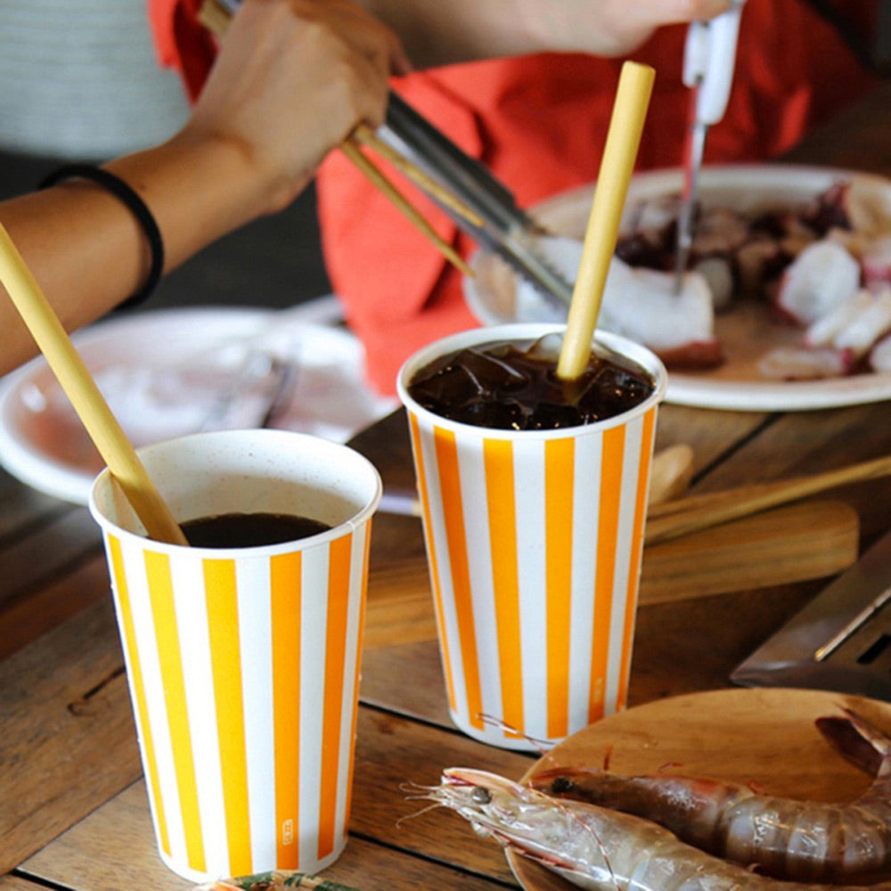 Image for two cups of soft drink with reusable bamboo drinking straws in them. There is also some other food shown placed next to them on a wooden surface.